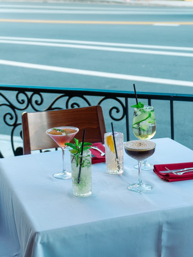 Five colorful cocktails on a white-clothed table on a restaurant patio overlooking a street.
