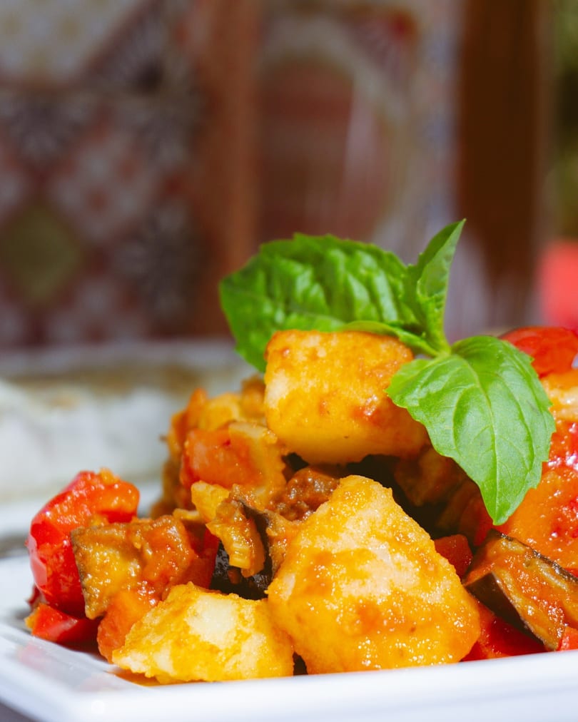 Close-up of golden fried potatoes, red peppers, and eggplant, garnished with fresh basil on a white plate.