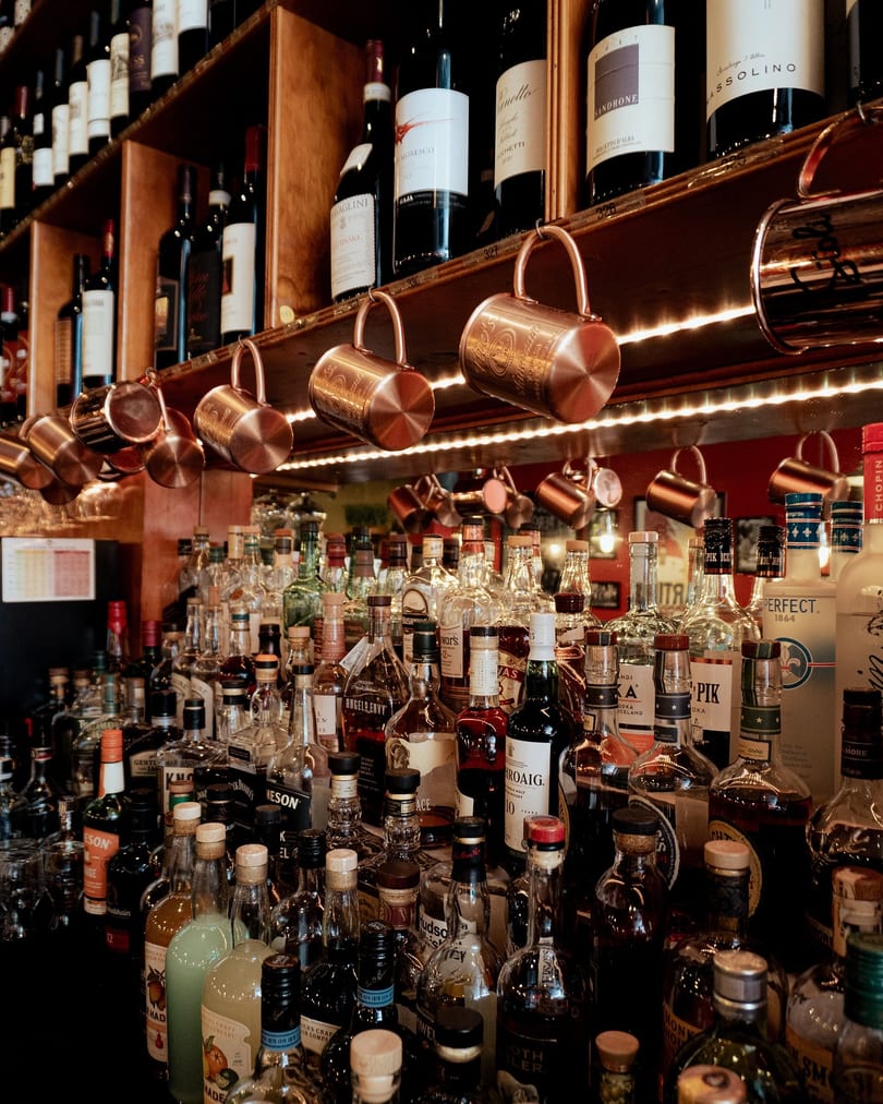A well-stocked bar with rows of liquor bottles, wine bottles on shelves, and copper mugs hanging above.