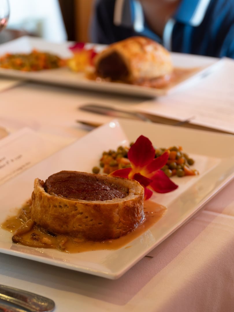 Beef Wellington with gravy, peas, carrots, and a red orchid on a white plate.